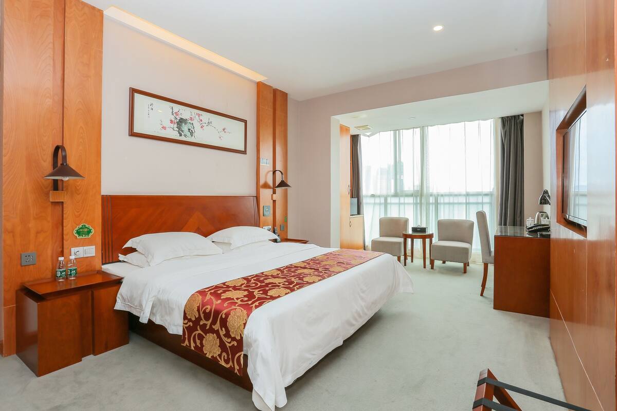 A hotel room with a large bed, a desk, a chair, a TV, and a window with a city view.