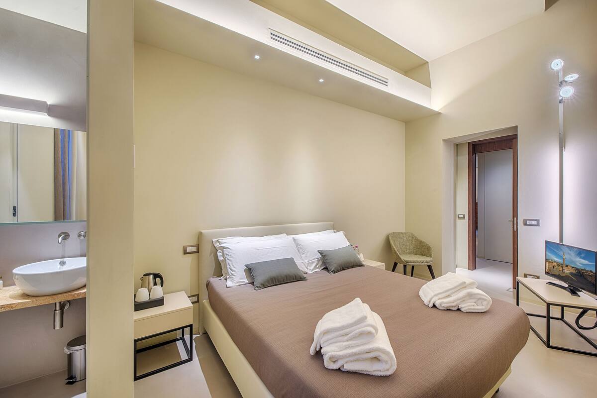 Een moderne hotelkamer met een groot bed, nachtkastje, televisie en een badkamer met wastafel en spiegel.