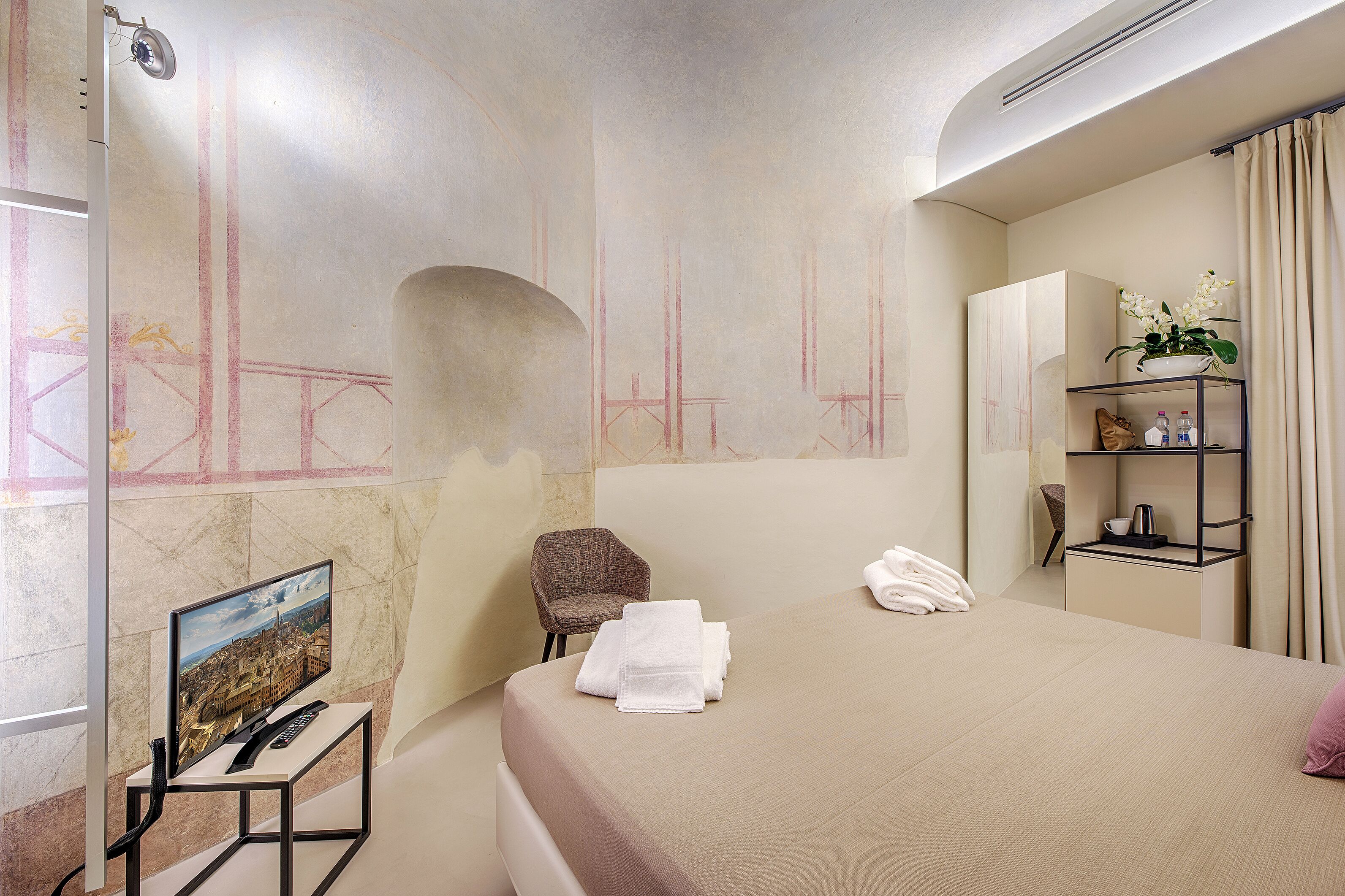 A hotel room with a bed, a chair, a TV, and a shelf with toiletries.