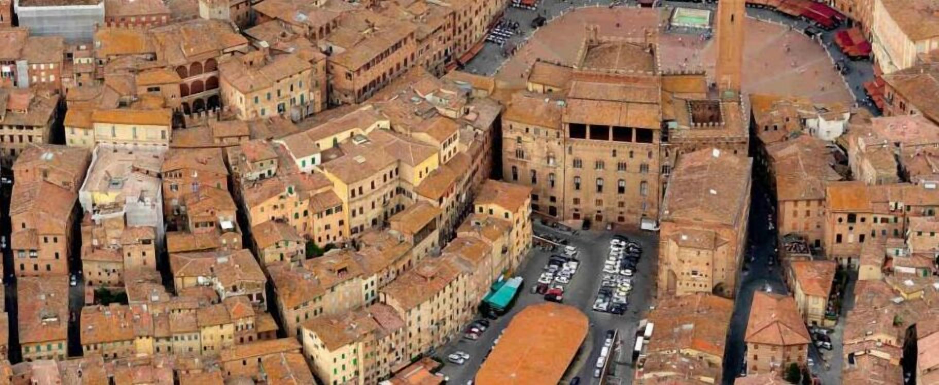 An aerial view of a densely packed urban area with closely built structures, narrow streets, and a central open space with parked vehicles.