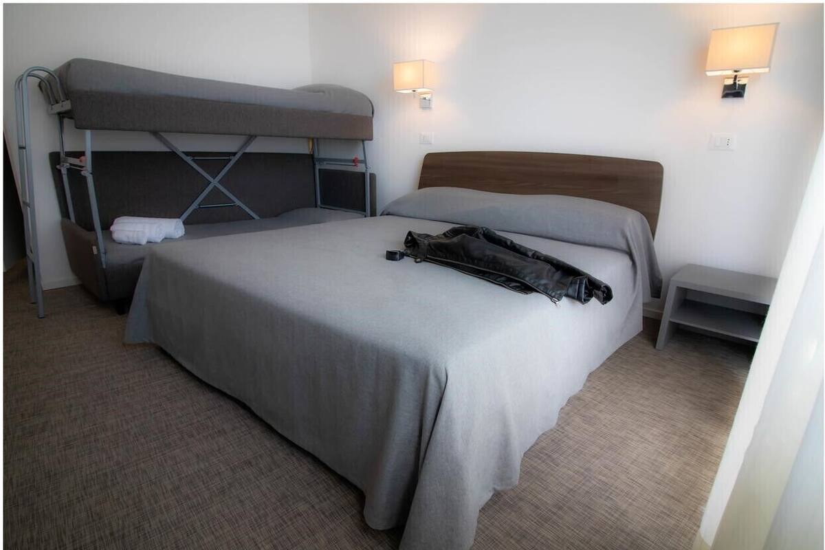 A single bed with a grey mattress and a wooden headboard. There is a grey bunk bed to the left, a small table, and two wall-mounted lamps.