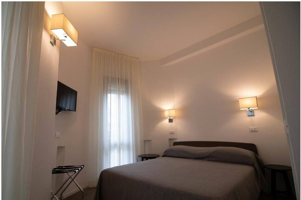 A hotel room with a bed, a TV mounted on the wall, two bedside tables, and a window with curtains.
