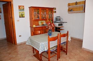 Zona pranzo con tovaglia a quadri, sedie in legno, un vaso con fiori, un mobile contenitore in legno con vetrate e un televisore su un supporto.