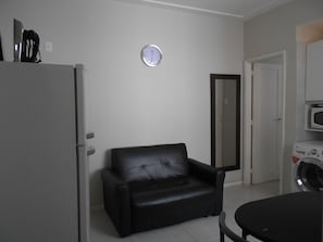 Ein kompakter Wohnraum mit Kühlschrank, schwarzem Ledersessel, Wanduhr, Tür und Waschmaschine.