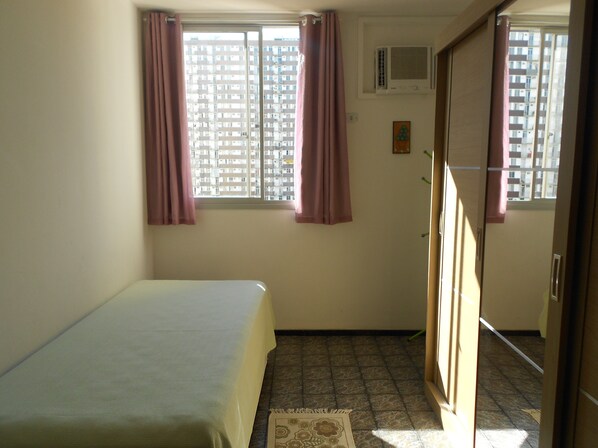 A small, sparsely furnished room with a bed, two windows with curtains, an air conditioning unit, and a door leading to another room.