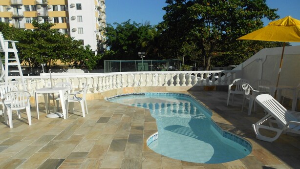Una piscina de borde curvo, rodeada de sillas de plástico blanco y un paraguas amarillo.