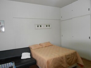 A single bed with a beige bedspread, a black sofa with a striped cushion, and a white towel on the sofa. There is a white wall with a small plant shelf and a door to the right. - Gohouse - Siqueira 1037 (Rio de Janeiro)