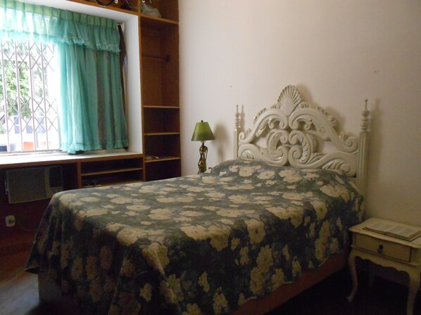 A bedroom with a bed, a nightstand, a lamp, a window with curtains, and a shelf unit.