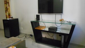 A modern living room with a flat-screen TV mounted on the wall, a glass coffee table, a wooden side table, and a black speaker. - Gohouse - Visconde 402 B (Rio de Janeiro)