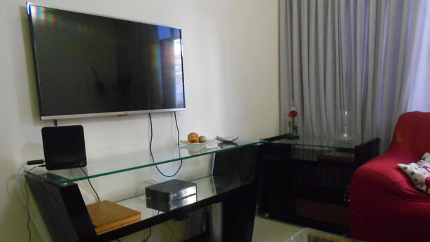 A living room with a flat-screen TV mounted on the wall, a glass coffee table, a red armchair, and a glass TV stand with electronic devices. - Gohouse - Visconde 402 B (Rio de Janeiro)