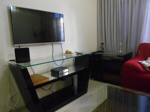 A living room with a flat-screen TV mounted on the wall, a glass coffee table, a red armchair, and a glass TV stand with electronic devices. - Gohouse - Visconde 402 B (Rio de Janeiro)