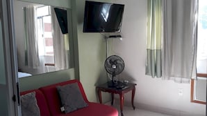 A room with a red sofa, a television mounted on the wall, a standing fan, and a window with curtains. - Gohouse - Siqueira 1417 (Rio de Janeiro)