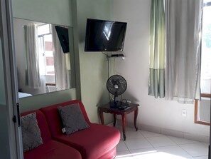 Cozy living space with a sofa, flat-screen TV, and standing fan, featuring a large mirror and sheer curtains.