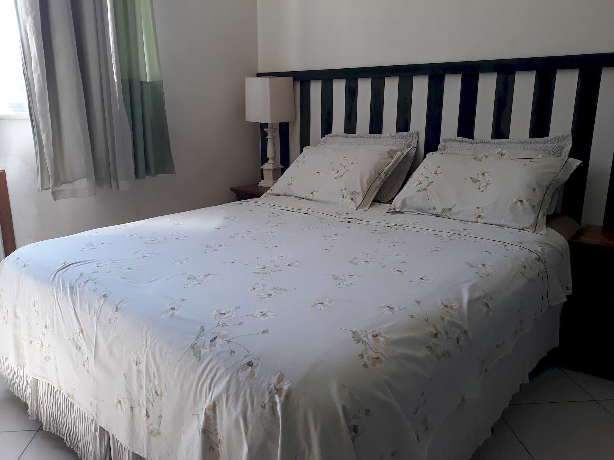 Ein ordentlich bezogenes Bett mit Blumenmuster auf der Bettwäsche, einem gestreiften Kopfteil und einer Nachttischlampe.