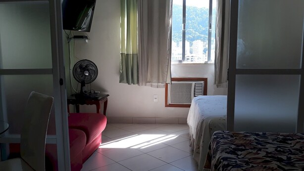 A hotel room with a bed, a chair, a small table with a fan, a TV mounted on the wall, and a window with curtains. - Gohouse - Siqueira 1417 (Rio de Janeiro)