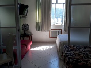 A hotel room with a bed, a chair, a small table with a fan, a TV mounted on the wall, and a window with curtains. - Gohouse - Siqueira 1417 (Rio de Janeiro)