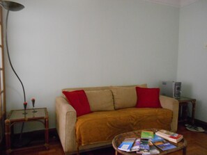 A living room with a beige sofa, red cushions, a small side table with a lamp, and a wicker coffee table with books. - Gohouse - Ferreira 76 (Rio de Janeiro)