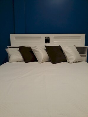 Cama com roupa de cama branca e duas almofadas, uma marrom e outra branca, em frente a uma parede azul.