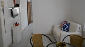 Une petite pièce propre, avec un canapé blanc, une table en verre, des chaises en osier, un téléviseur posé sur une étagère et une porte donnant sur une autre pièce.