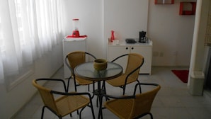 Un petit coin repas avec une table ronde et quatre chaises en osier. Un réfrigérateur blanc, surmonté d’un appareil rouge, est adossé au mur.