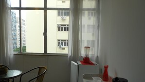 Une petite pièce avec une table, deux chaises en osier, un radiateur blanc, un objet rouge posé dessus, et une vue sur les bâtiments à l’extérieur.