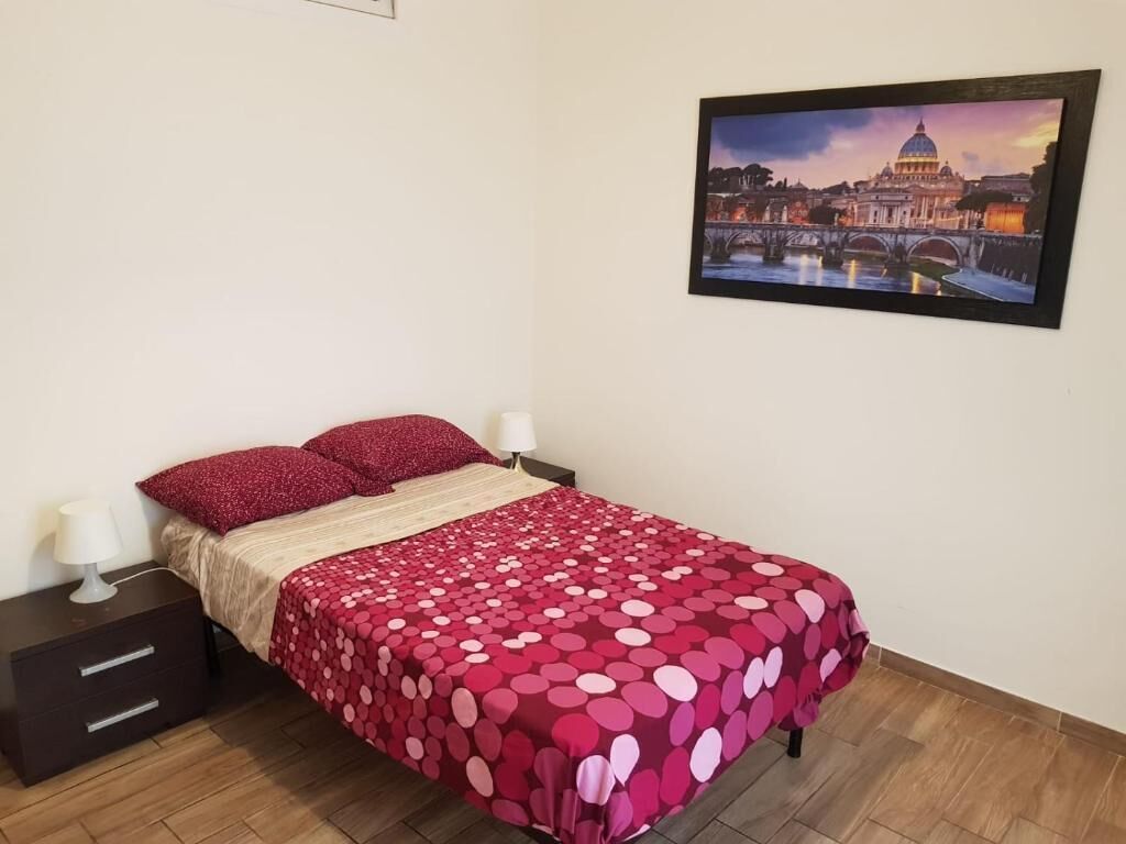 A neatly made bed with a red polka dot bedspread, a dark wooden nightstand with a lamp, and a framed picture of a cityscape on the wall.
