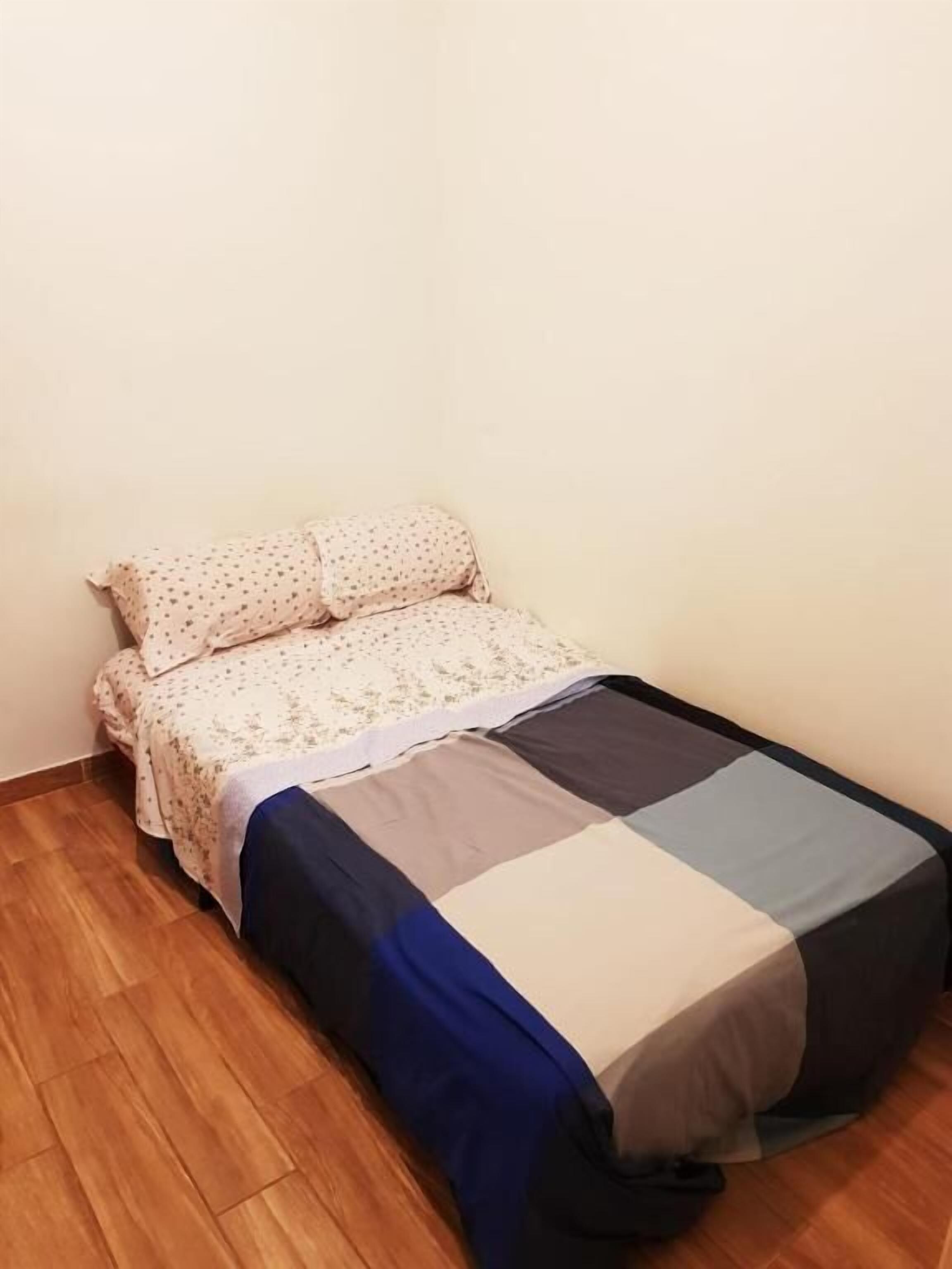 Ein kleines Zimmer mit einem Einzelbett, Holzboden, einem Bett mit floralem Überwurf und Kissen sowie einer schlichten Wand.