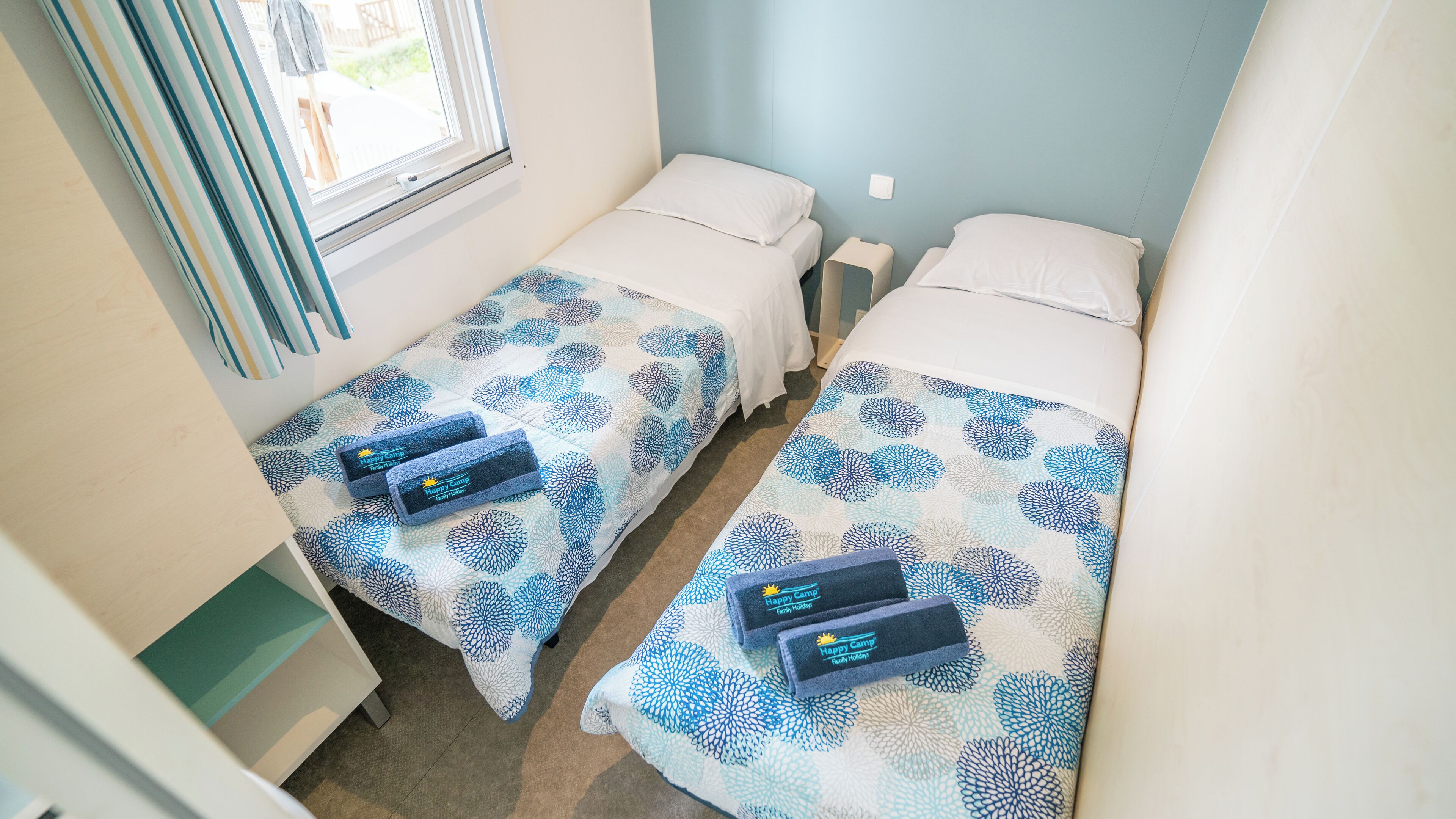 Habitación pequeña y limpia con dos camas individuales, cada una con una almohada a juego y una toalla con el logotipo 'Happy Camp Family Holidays'.