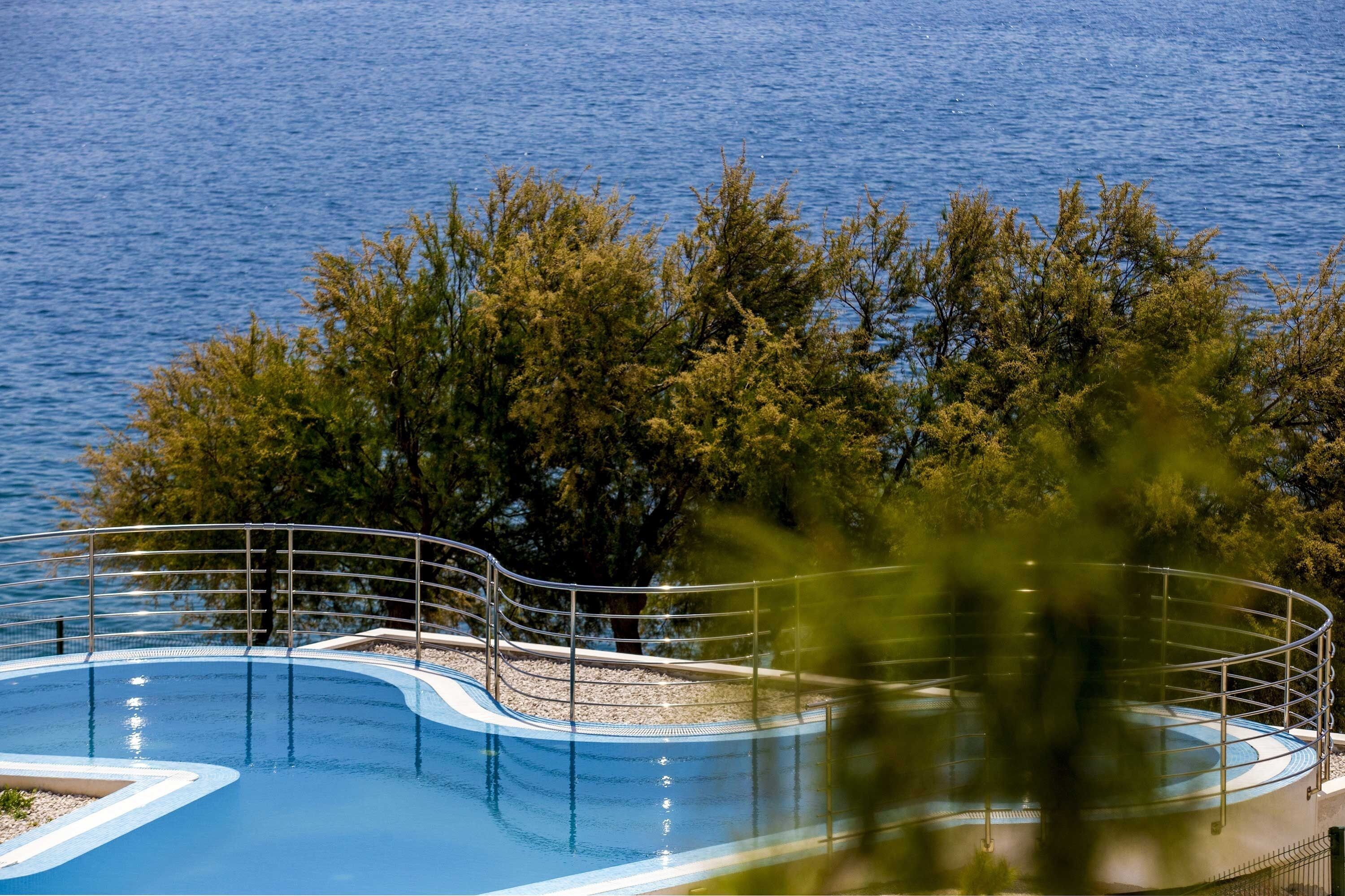 Une piscine entourée d’arbres, dotée d’une rambarde en métal et offrant une vue sur une étendue d’eau.