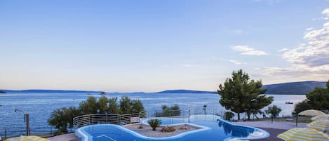 Área da piscina com vista para o mar, rodeada por árvores e guarda-sóis.