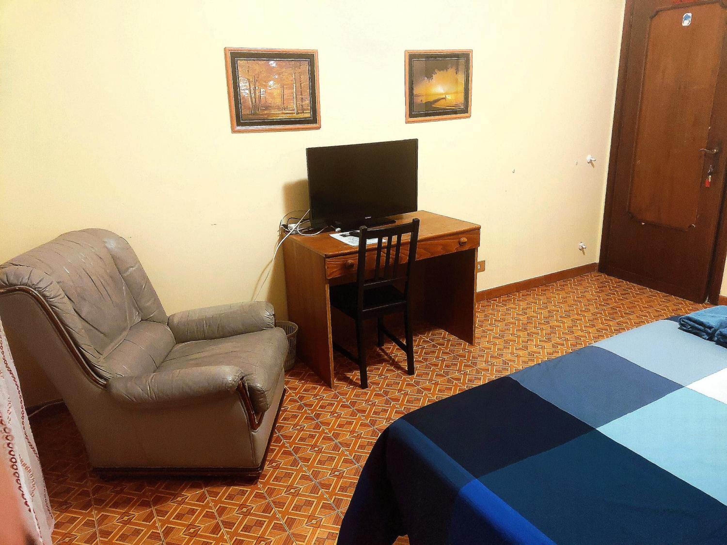 A room with a bed, a desk with a computer, a chair, and two framed pictures on the wall.