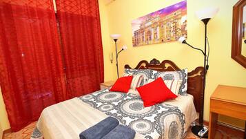 A hotel room with a bed, red curtains, a painting on the wall, a lamp, and a wooden nightstand.