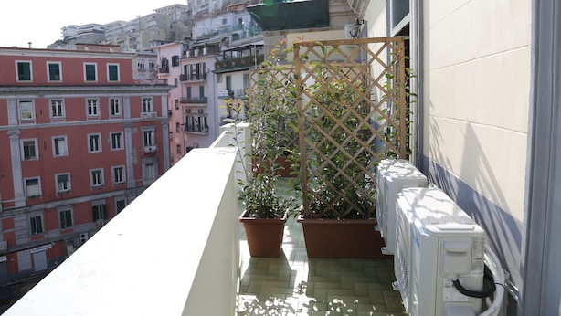 A balcony with potted plants, a white air conditioning unit, and a view of adjacent buildings. - Sun&TheCity (Naples)