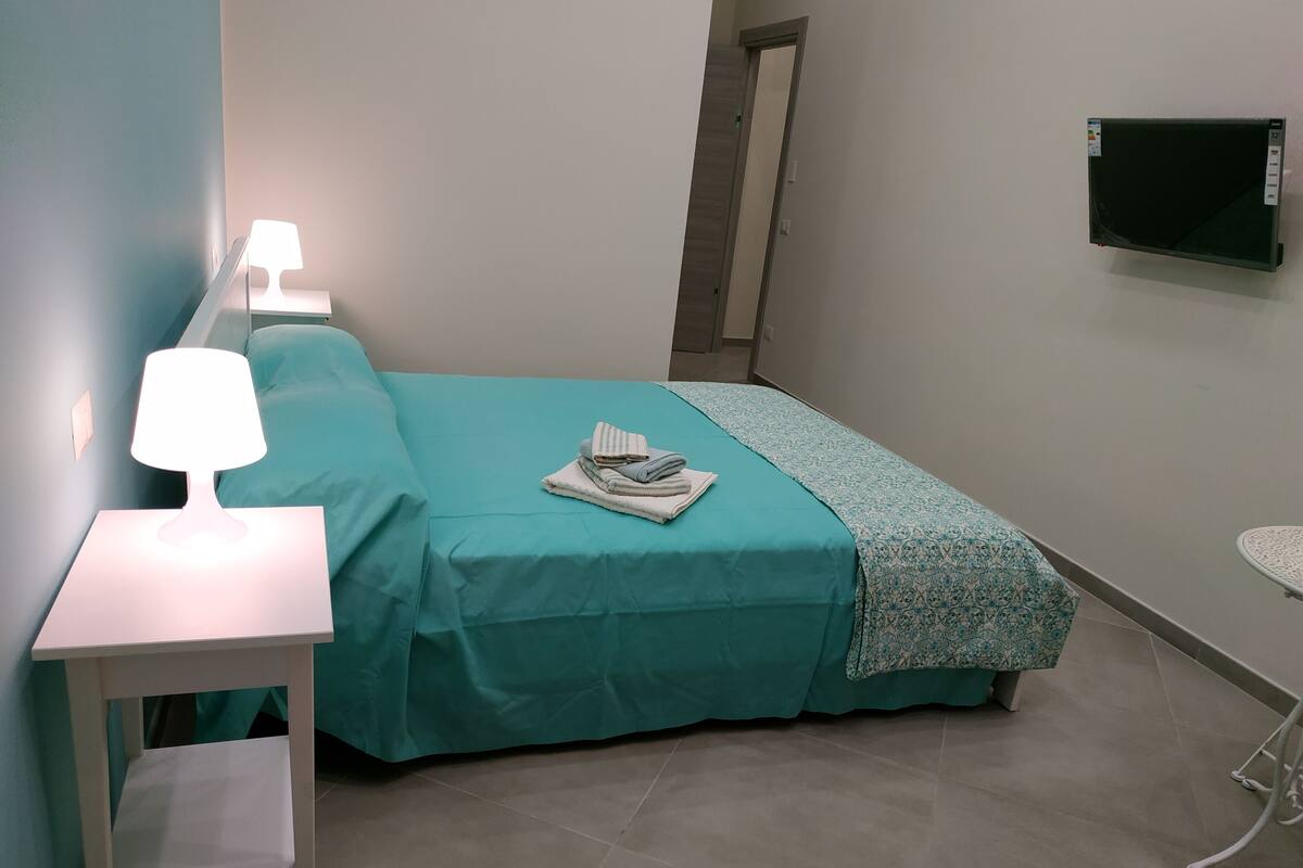 A small, neatly arranged bedroom with a bed, a bedside table with a lamp, a TV on the wall, and a small table with a chair.