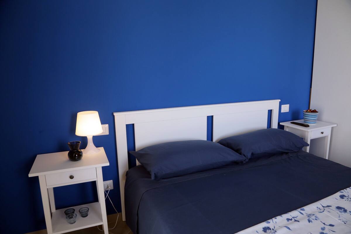 A bedroom with a bed, a nightstand with a lamp, and a small table with a remote control.