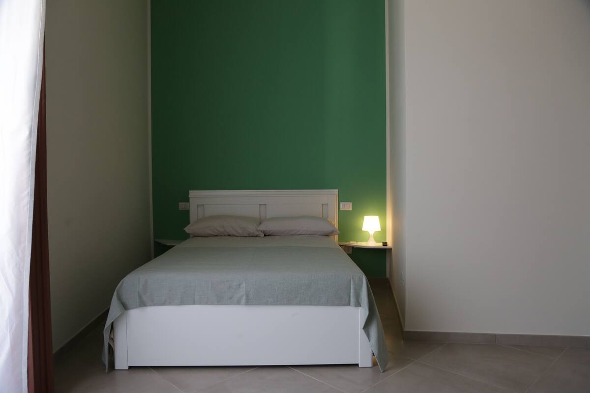 A single bed with a white headboard in a room with green walls and a white nightstand with a lamp.