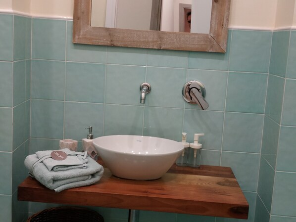 Une salle de bain avec un lavabo blanc, deux robinets et un miroir au-dessus. Des serviettes sont pliées sur une étagère en bois, et une petite bouteille est posée sur le lavabo.