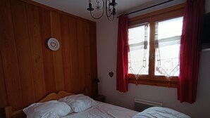 A bedroom with a wooden headboard, a bed with white bedding, a bedside table with a lamp, a window with red curtains, and a wall clock. - Cozy apartment in Montgenèvre - Close to slopes - South-facing terrace (Montgenèvre)