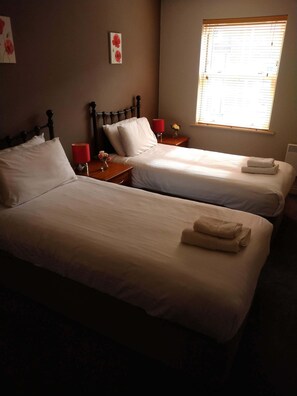 A hotel room with two single beds, a nightstand with a red lamp, and a window with blinds. - Victoria Cloisters Apartments (York)