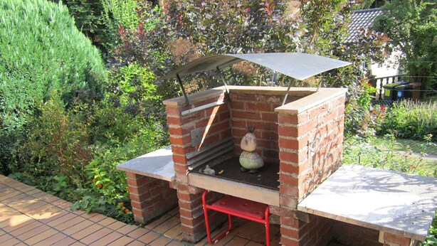 A brick barbecue grill with a chimney, a small statue, and a tiled patio area. - Holiday house without everyday noise (Altomünster)