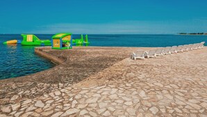 A beachside area with a stone walkway, inflatable play structures, and white lounge chairs. - Happy Camp in Camping Park Umag (Karigador)