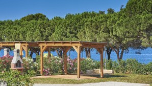 A wooden pergola with a stone fireplace, surrounded by a garden with various plants and flowers, overlooking a body of water. - Happy Camp in Camping Park Umag (Karigador)