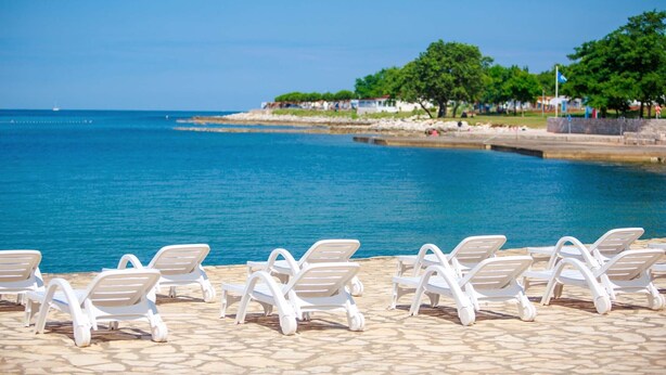 A beachside area with lounge chairs facing the sea, a stone pathway, and a distant shoreline with trees and buildings. - Happy Camp in Camping Park Umag (Karigador)