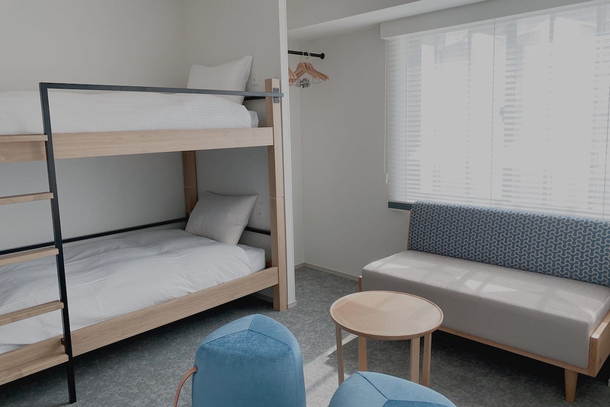 A bunk bed with a small table and a sofa in a room with a window.