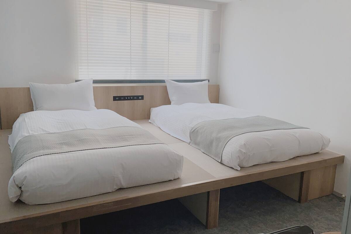 A bedroom with two beds, a wooden headboard, a window with blinds, and a control panel on the wall.