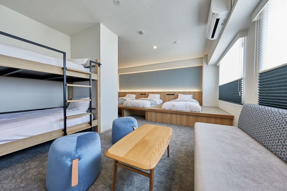 A hotel room with bunk beds, a sofa, a small table, and a window with blinds.