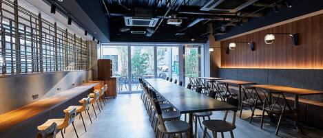 A modern dining area with long tables and chairs, a wooden accent wall, and a view through large windows.