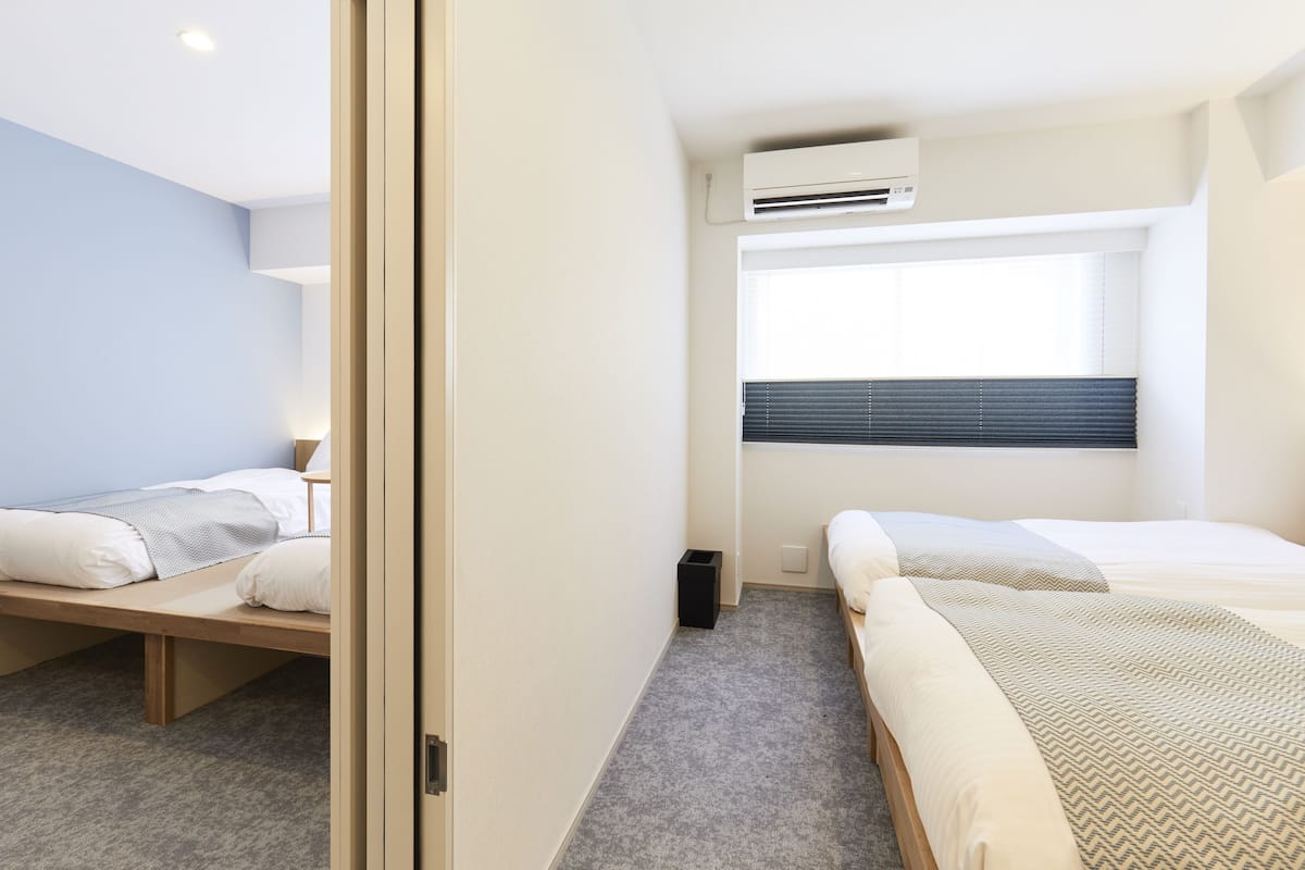 A modern hotel room with two single beds, a small window with blinds, an air conditioning unit, and a carpeted floor.