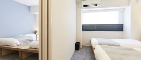 A modern hotel room with two single beds, a small window with blinds, an air conditioning unit, and a carpeted floor.