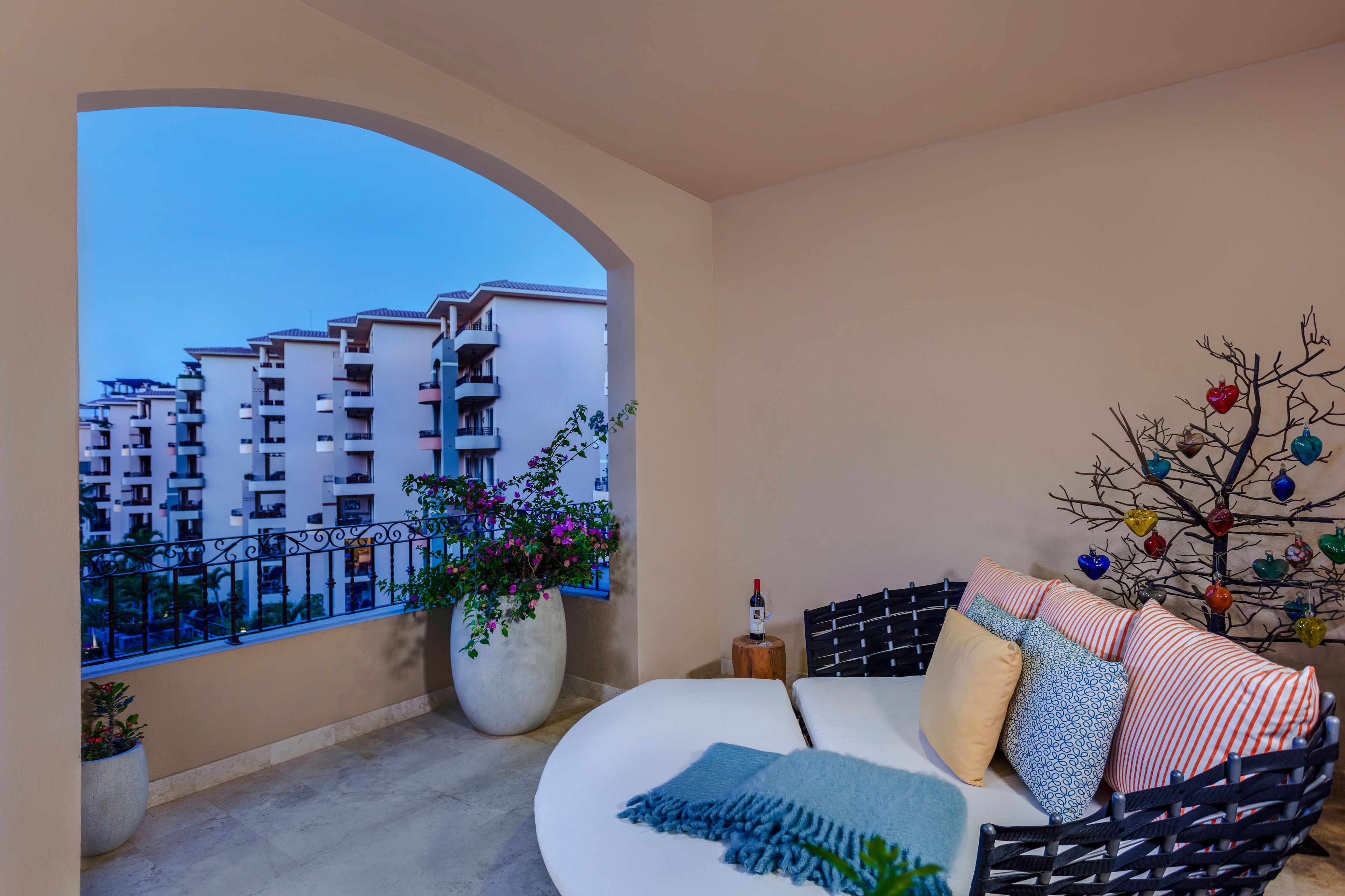 Ein Balkon mit Blick auf hohe Gebäude, einem großen Topfpflanzen, einem Rattansofa mit Kissen und einem Zierbaum.